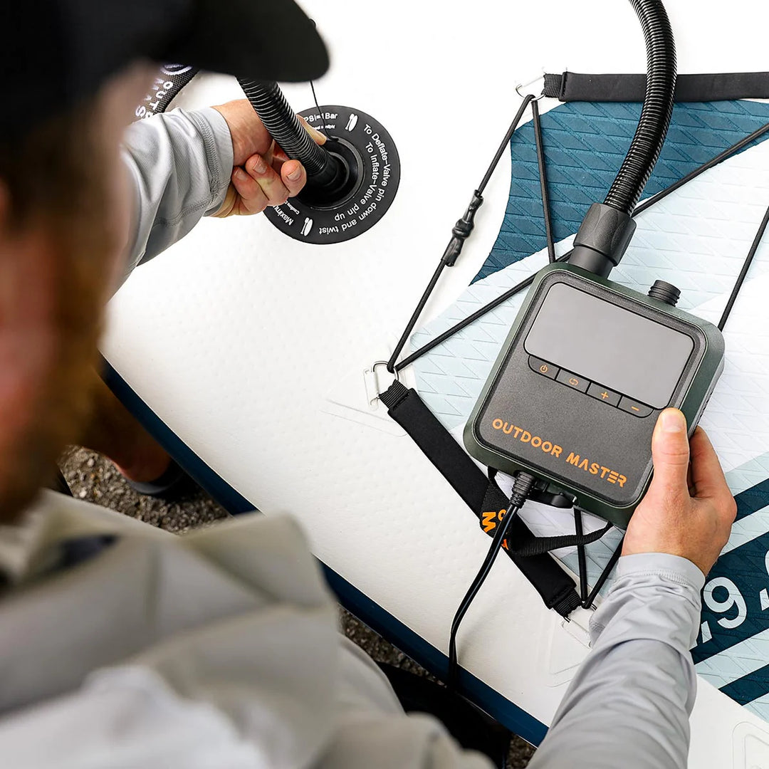 Person using a SUP pump labelled 'Outdoor Master' on a paddleboard with a focus on the device.
