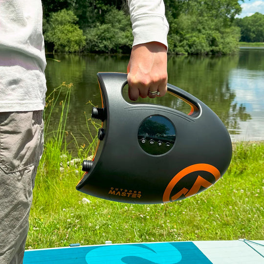 Person holding a black and orange portable electric SUP pump by a lake