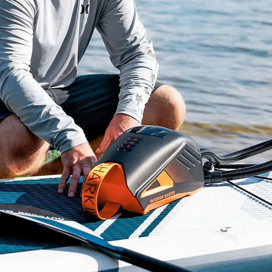 Person inflating a stand-up paddleboard with an Outdoor Master pump on a body of water.