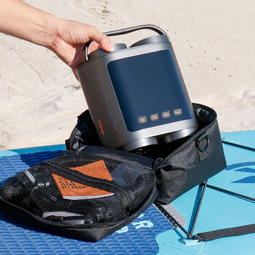 Hand placing a portable SUP pump into a black carrying case on a blue iSUP on a beach.