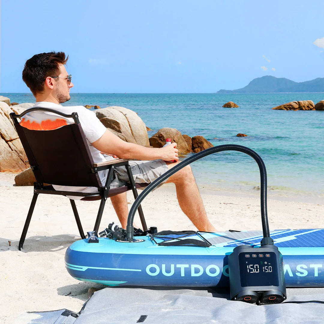 Man sitting on a beach with an inflatable paddleboard and electric pump.