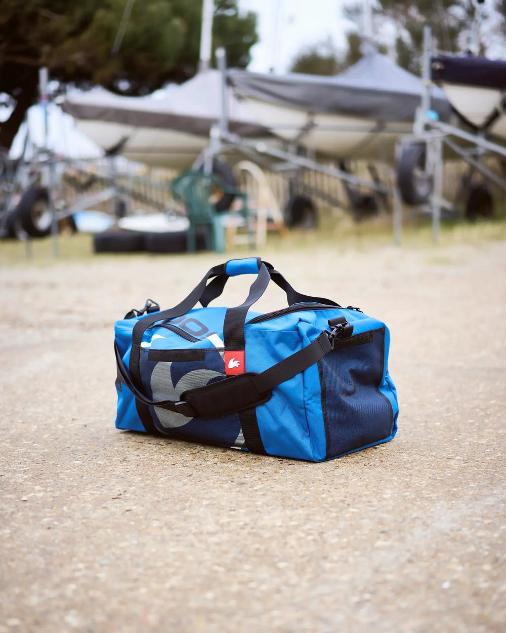 Blue duffel bag with a red logo on a gravel surface with blurred background.