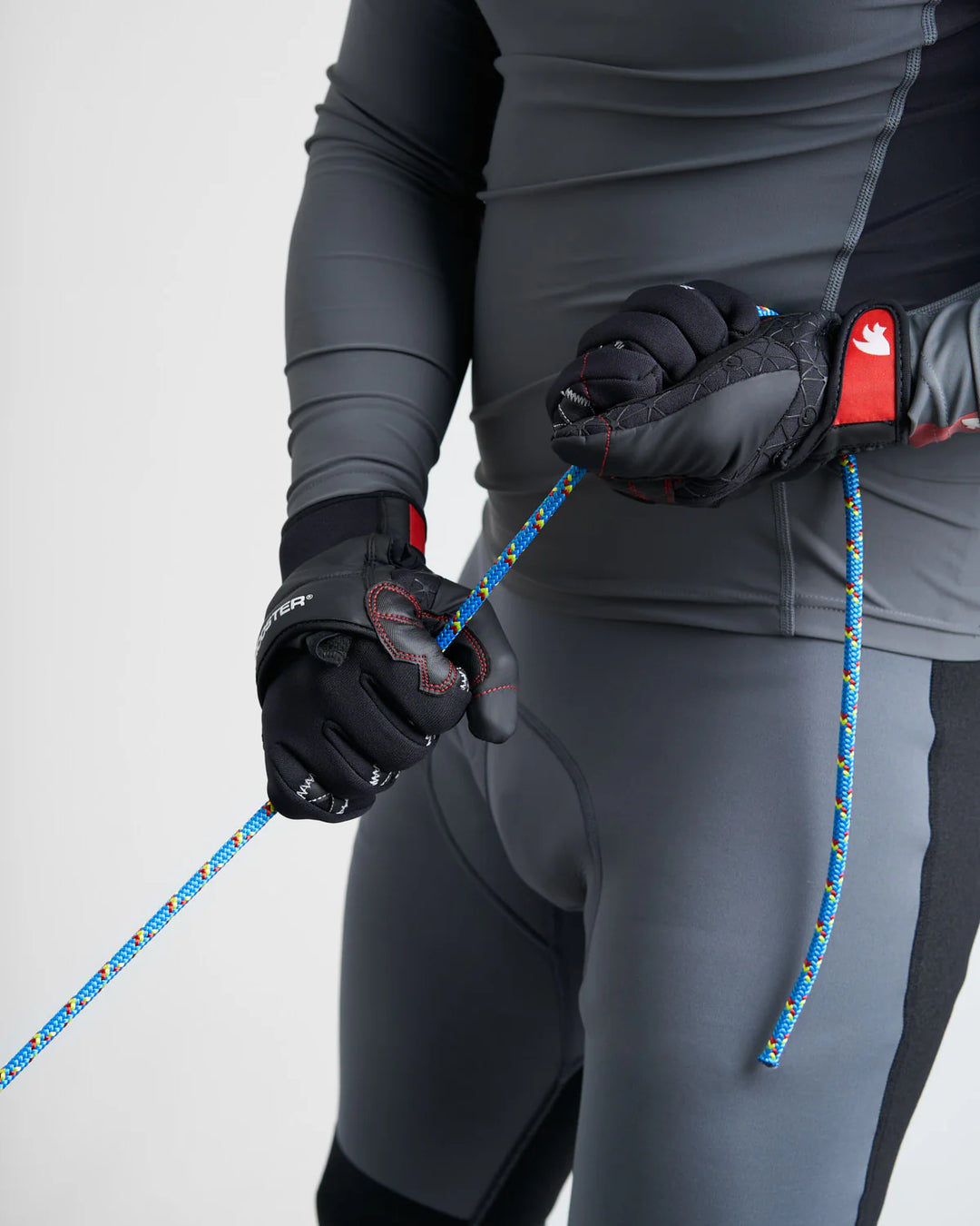 Person wearing black sailing gloves and grey pants holding a blue rope against a white background.