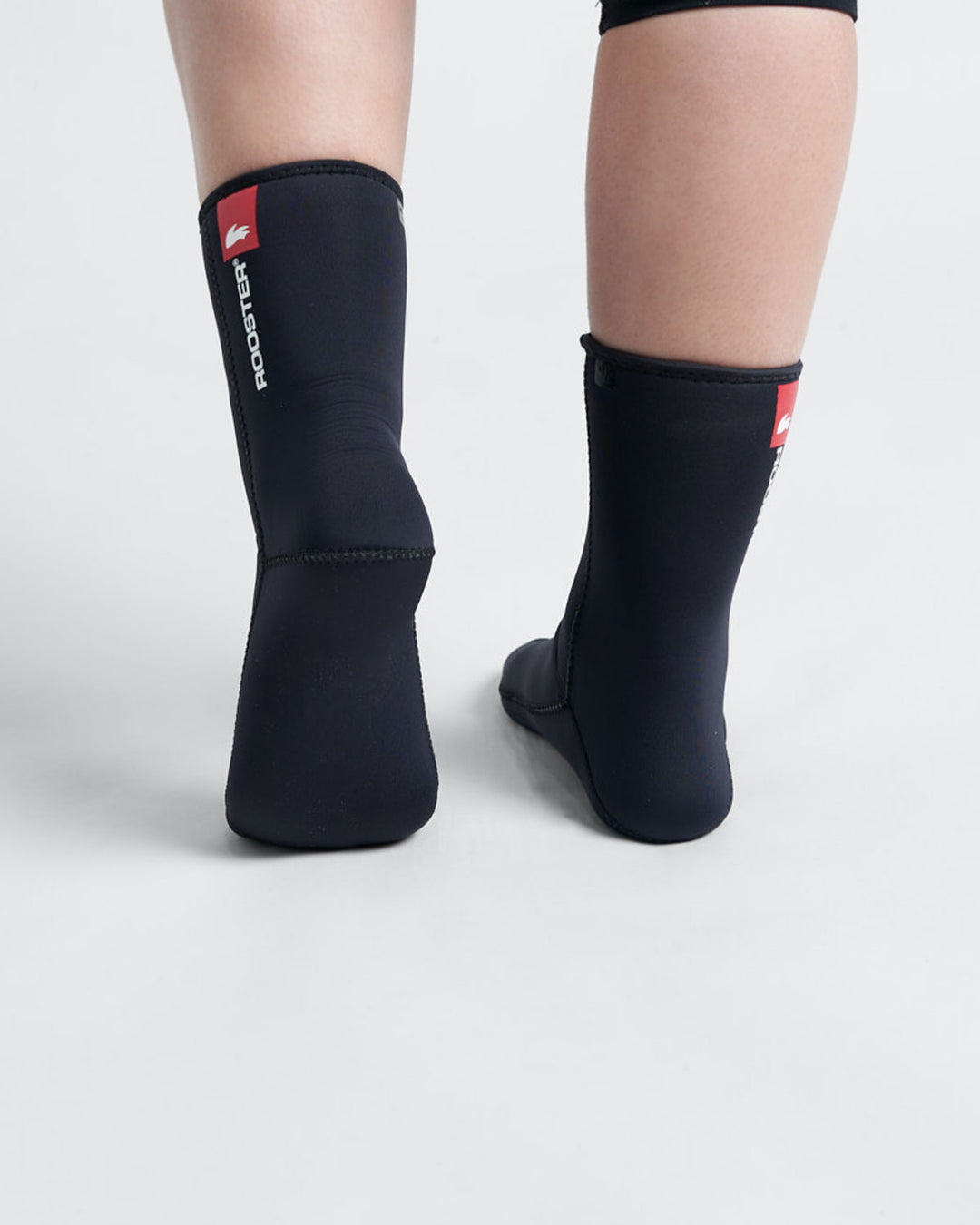 Studio image of the heals of a model wearing black 4mm thick neoprene socks with a white and red Rooster logo.