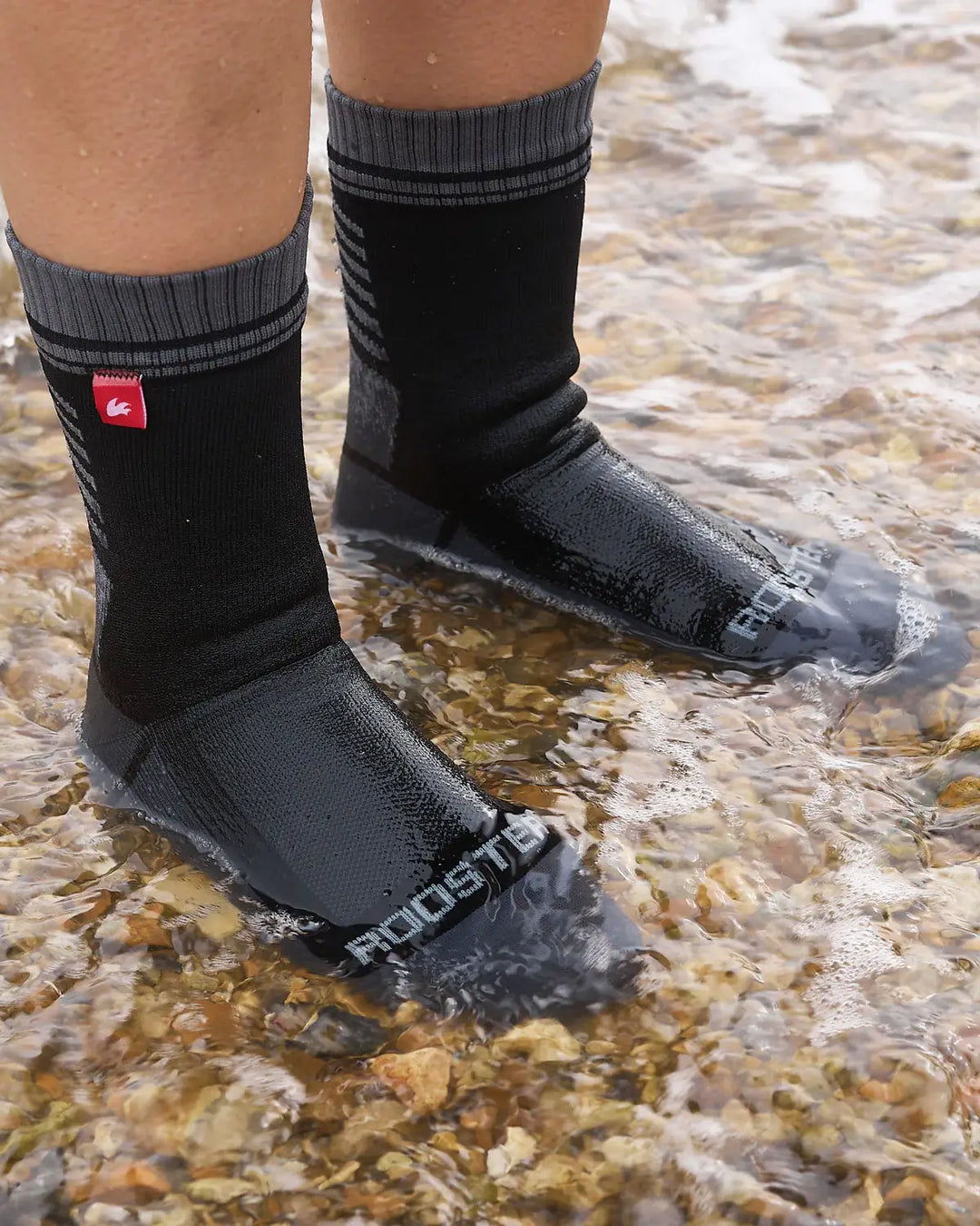 Grey and black striped waterproof sock with a red logo tag on, stood in clear water.