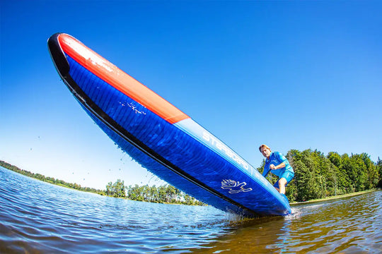 Person on a blue and red Starboard All Star Airline Inflatable 
Paddleboard on a lake with trees in the background