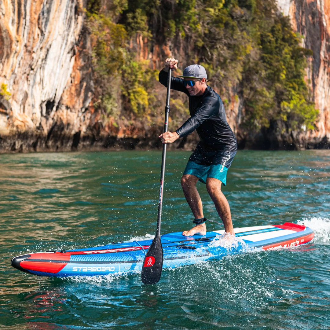 Image of a male paddleboarder on a blue Starboard All Star Airline 14'0 x 28 x 6, paddling in front of cliffs.
